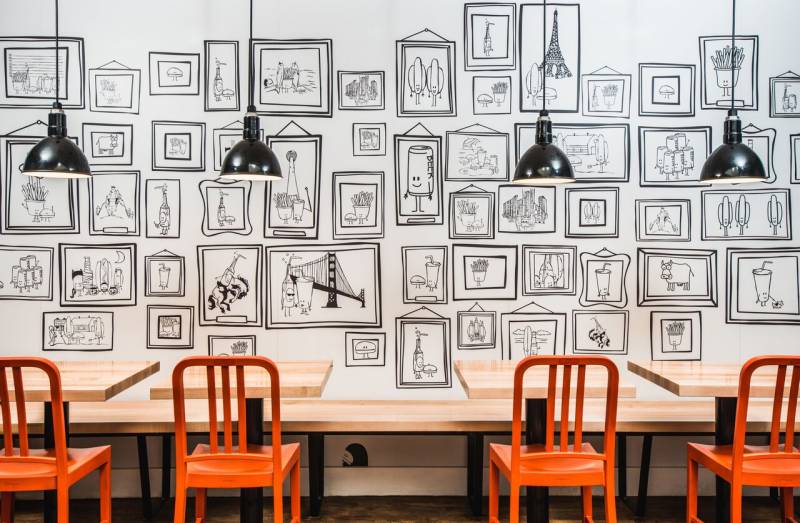 Wall with black cartoons across in front of tables with orange chairs 