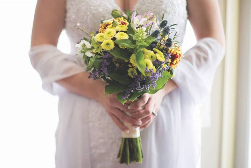 bride with white off the shoulder dress holding Sunflower bouquet with delphinium accents 