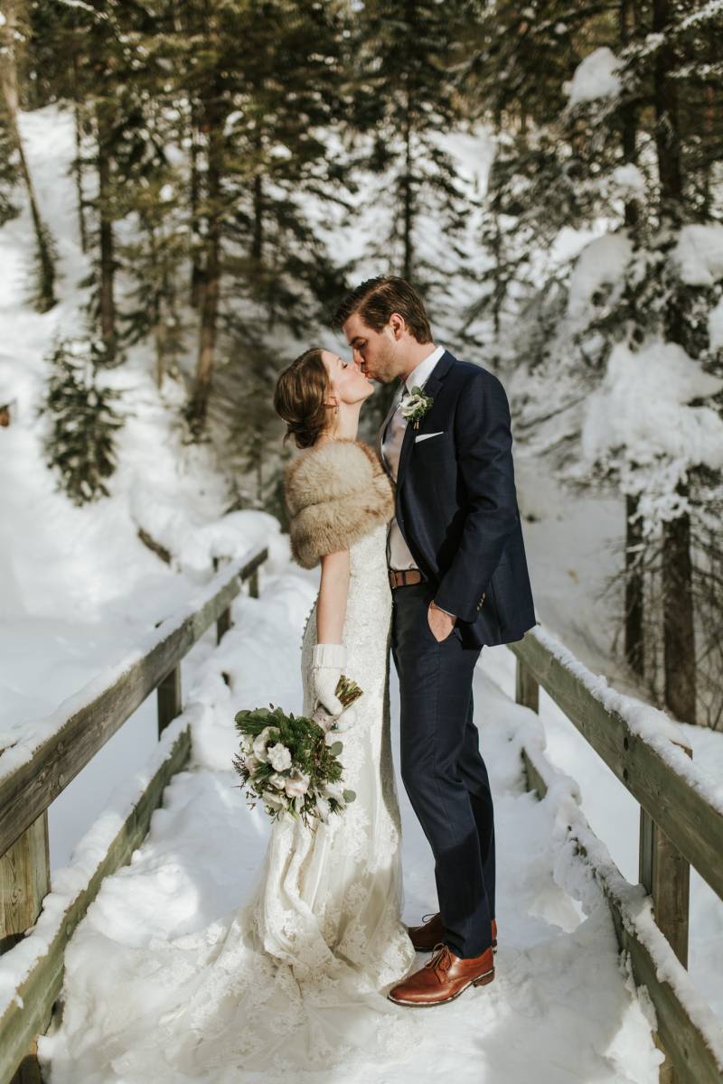 Bride and groom kiss on snowy wooden bridge 