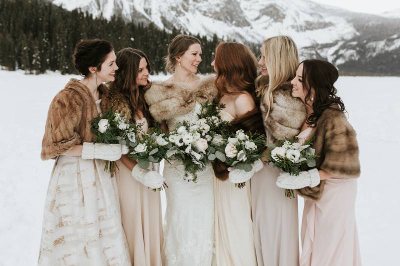 Bride and bridesmaids stand in snowy field wearing brown shawls holding white bouquets