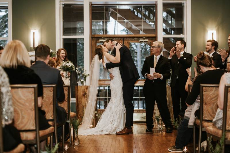Bride and groom kiss while guests clap on snowy cabin background