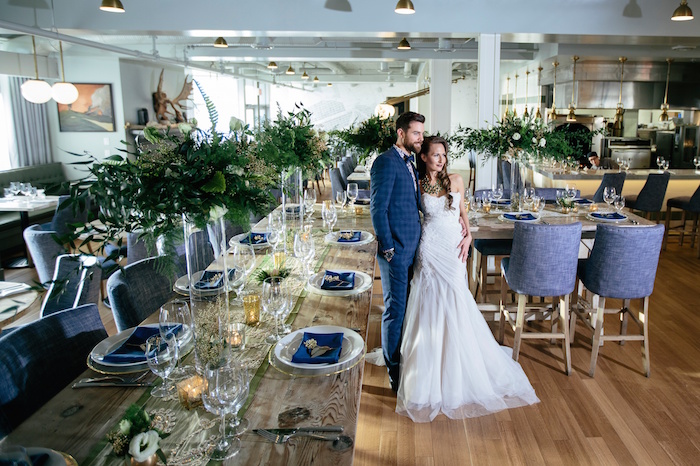 Man and woman stand embraced at edge of long wooden table with blue place setting and large green centerpiece 