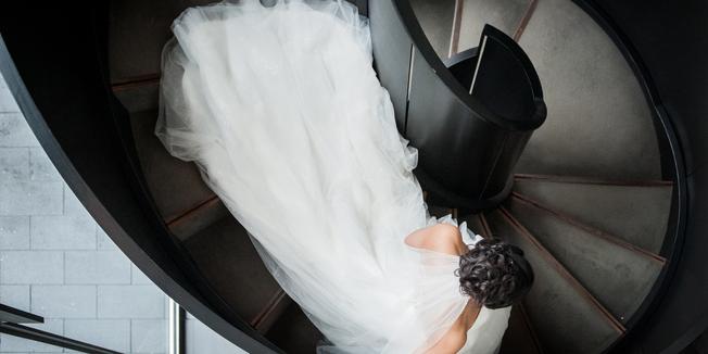 Woman in large white wedding dress and veil walks down winding black staircase from above