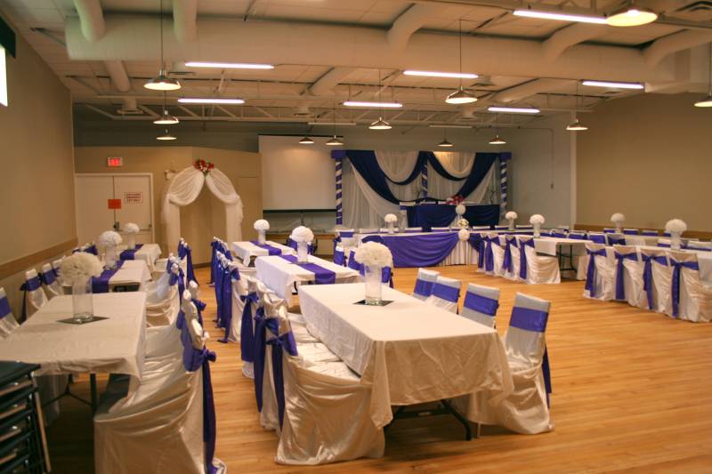 wooden floored room with white and purple decorated tables in front of white fabric wedding arch 