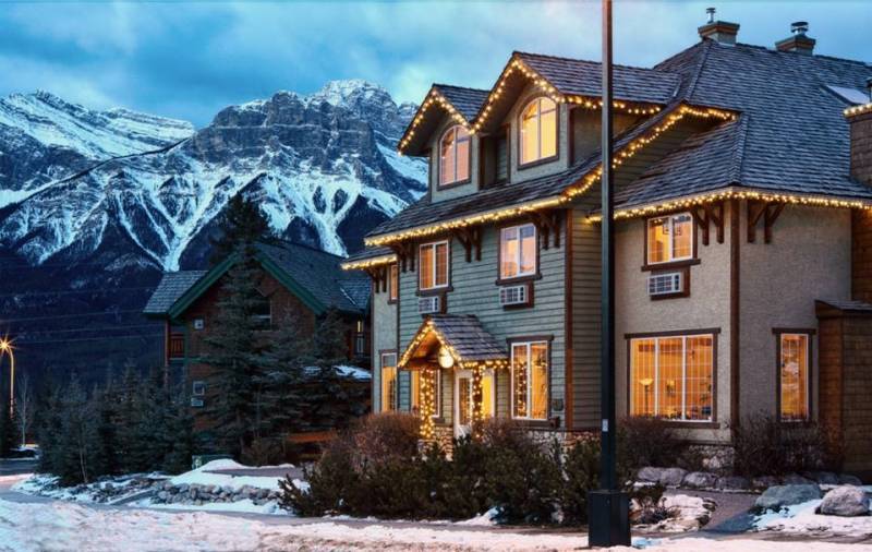 Large house with Christmas lights around edge in front of snowy rocky mountains at dusk 