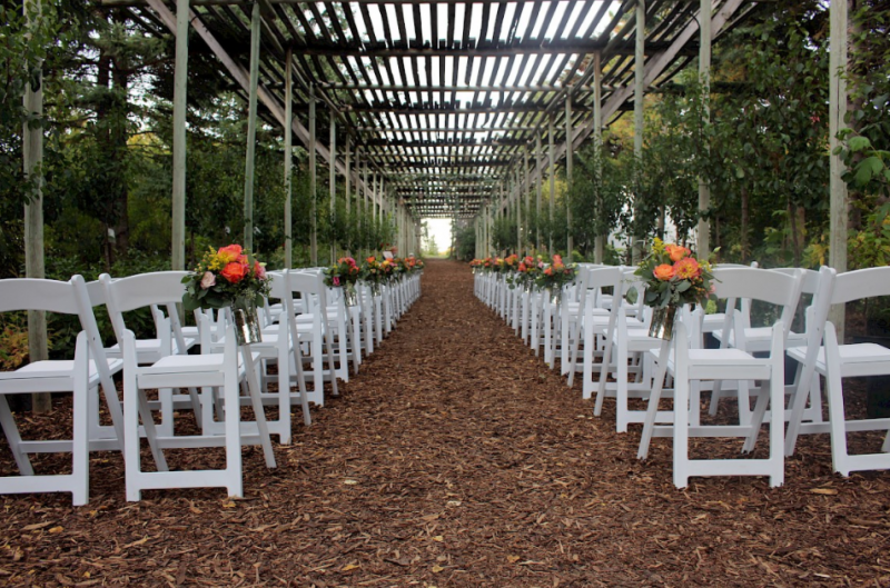 Aisle of white chairs and orange floral accents under slatted wooden overhead walkway 