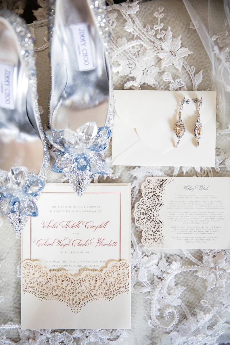 Wedding invitation flat lay on lace fabric beside silver crystal heels