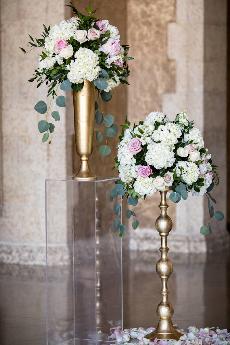 White floral arrangements set on clear pedestals with pedals surrounding  