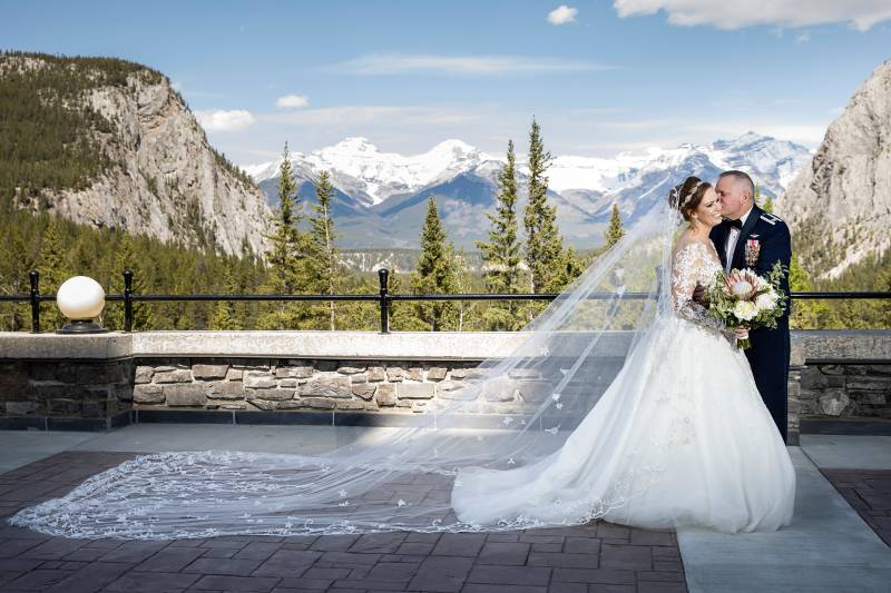 Bride stands sideways in large flowing veil while groom kisses cheek 