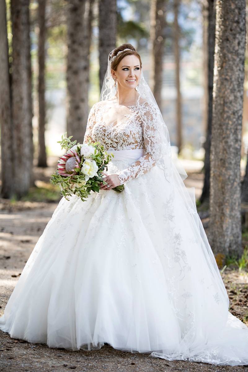 Bride standing in white dress and veil in wooded area smiling holding white bouquet 