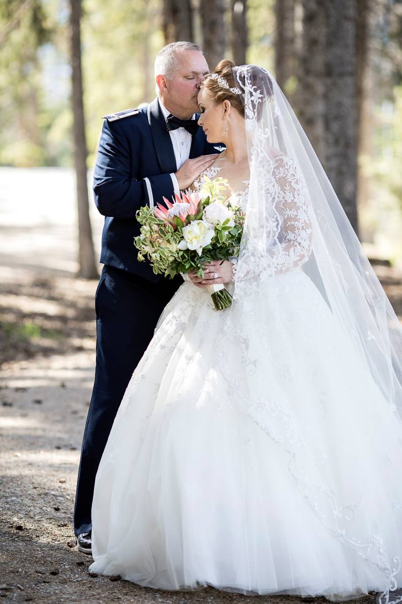 Groom kisses brides forehead holding bouquet in wooded area