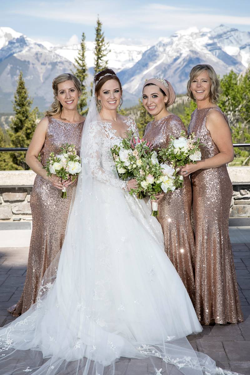 Bride stands with bridesmaids holding white bouquets backing rocky mountains 