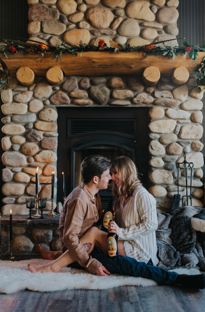 Woman in dress shirt sits in mans lap facing holding two black bottles in front of stone fireplace