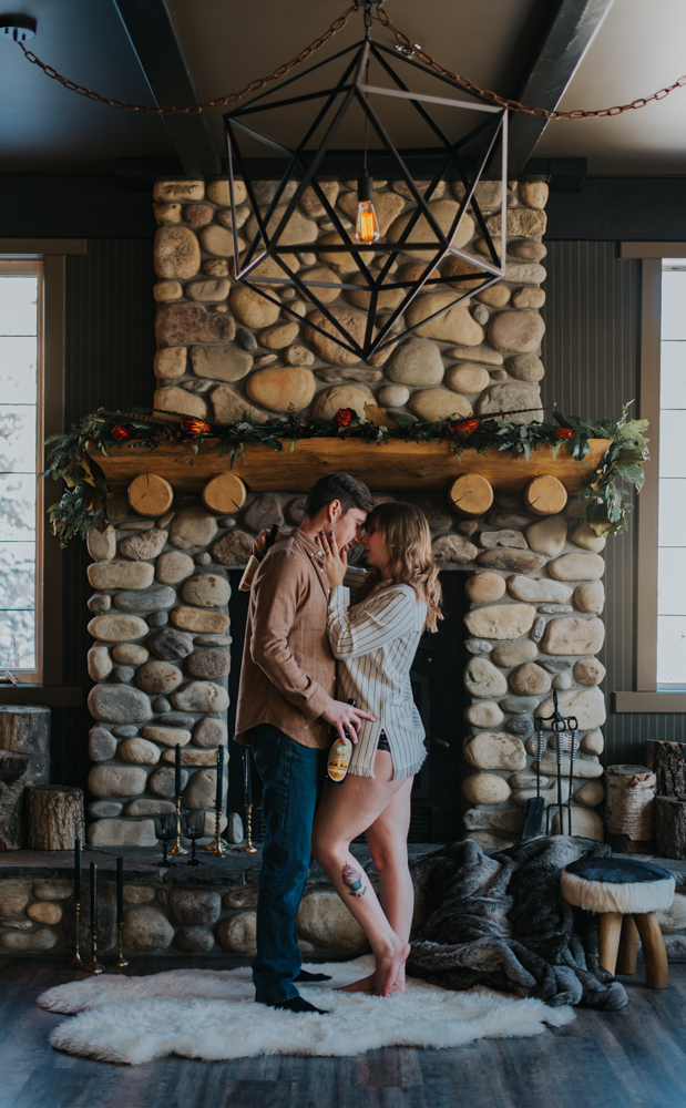 Man and woman stand embraced under geometric chandelier and stone fireplace 