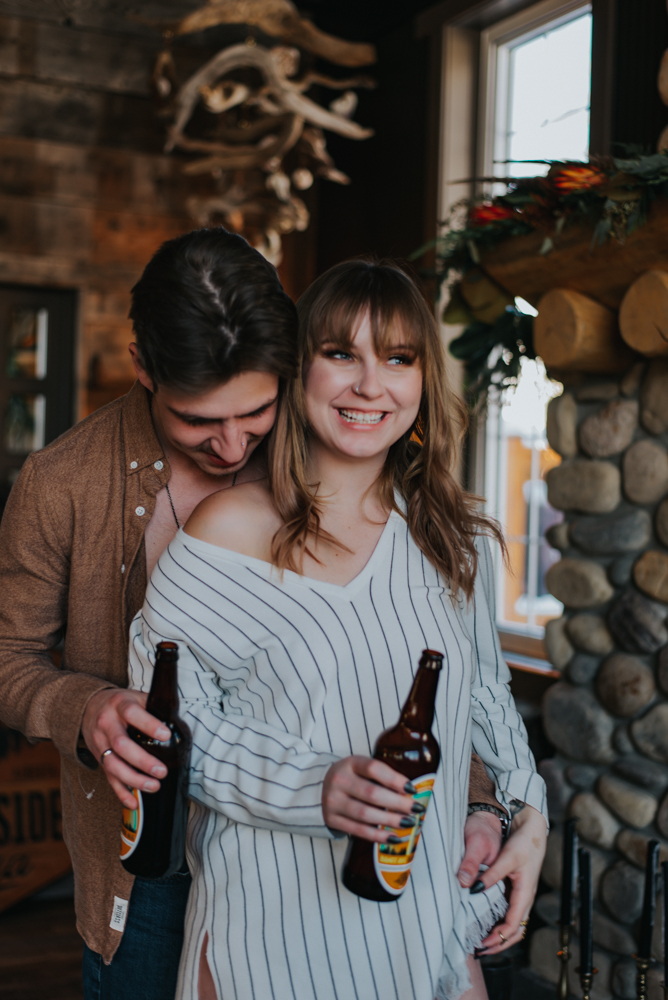 Man leans over woman's shoulder smiling holding glass bottle 