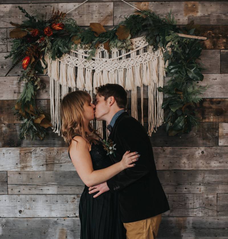 Man and woman kiss embraced in front of macramé wall 