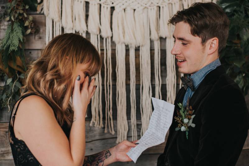 Woman in black wedding dress wipes eye while reading vows in front of macramé wall 