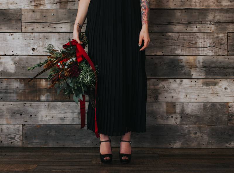 Woman in black dress holds crimson and rust bouquet to side with red ribbon hanging