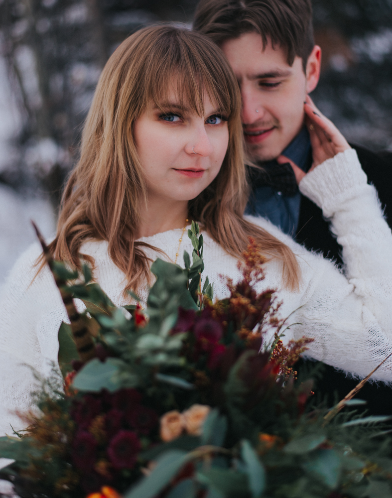Man looks over woman's shoulder as she touches chin bolding crimson bouquet in front 