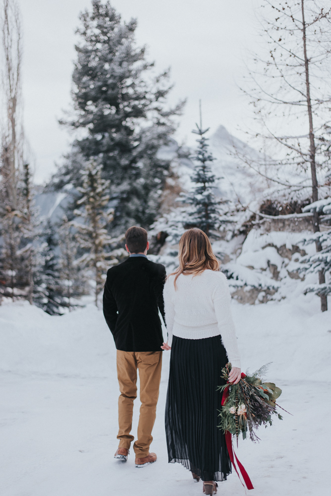 Man and woman walk holding hands and bouquet with red ribbon on snowy pathway 