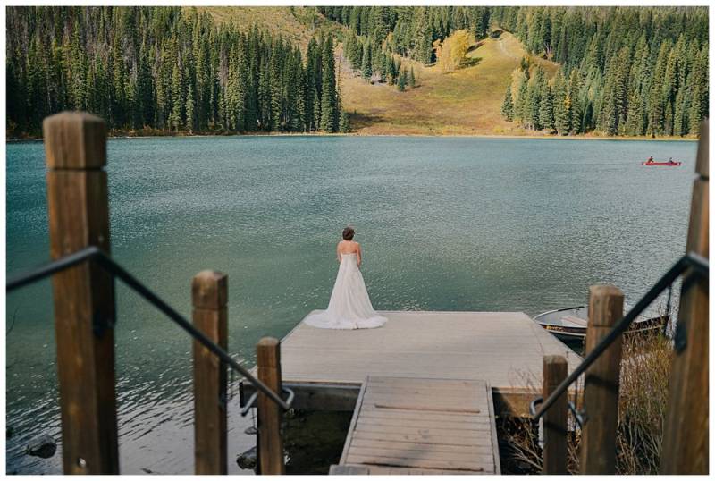 Bride stands at the edge of dock facing blue lake in white dress