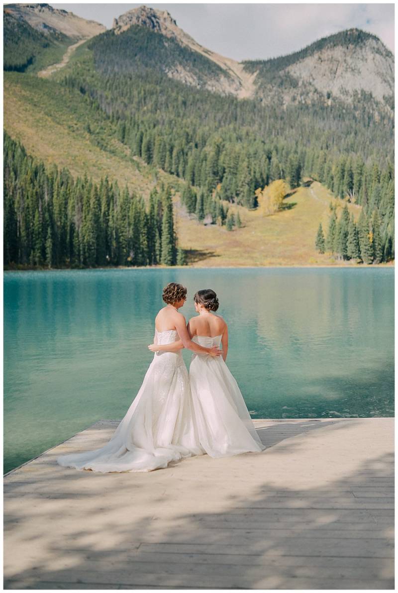 Brides standing in white dresses embracing facing blue lake on dock