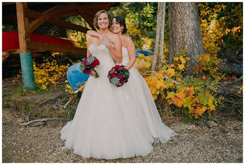 Brides wearing white shoulder-less dresses embracing holding red bouquets on forested backdrop