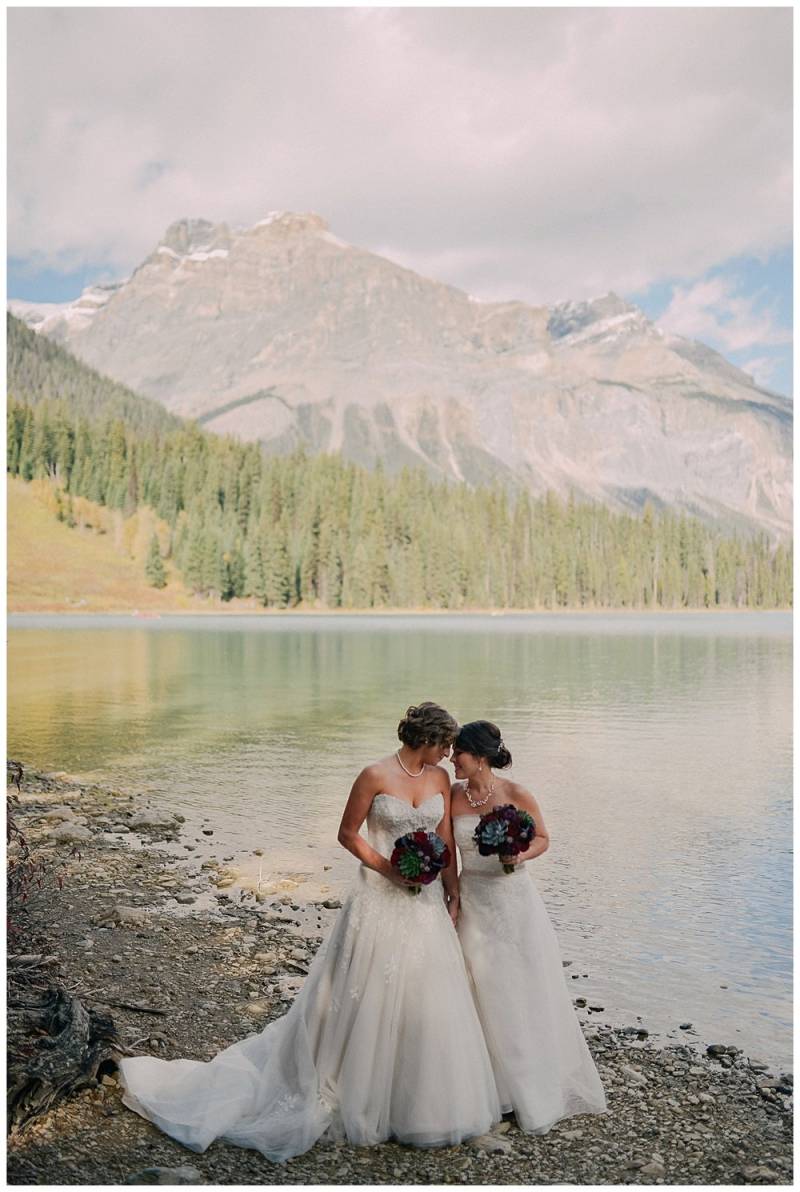 Brides wearing white dresses at the edge of lake on mountain backdrop