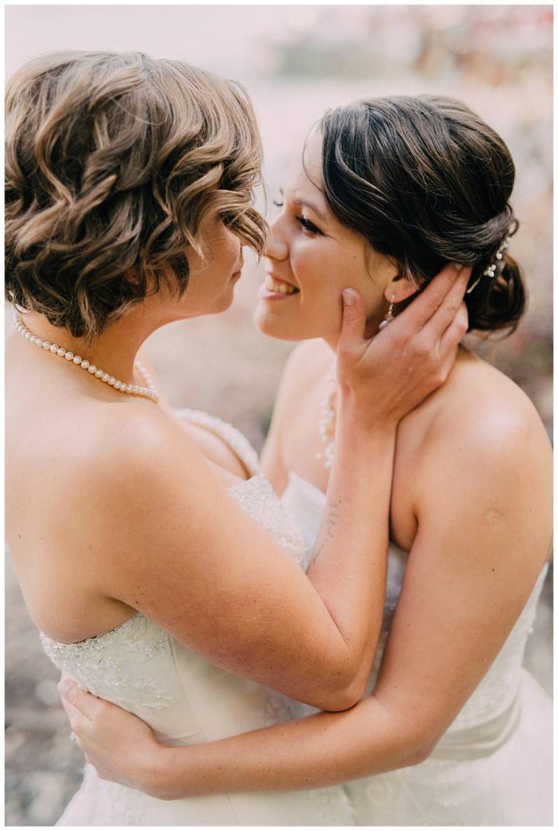 Brides embrace looking at each other in white lace dresses and pearl necklaces