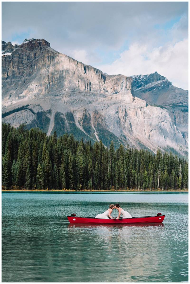 Brides in white dresses kiss on red canoe on lake with mountain background