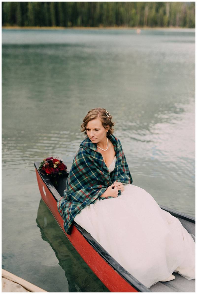 Bride wearing green plaid shawl and white dress sitting in red canoe on lake
