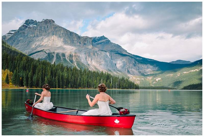 Brides in white dresses paddle red canoe on green lake water on mountain and forest background