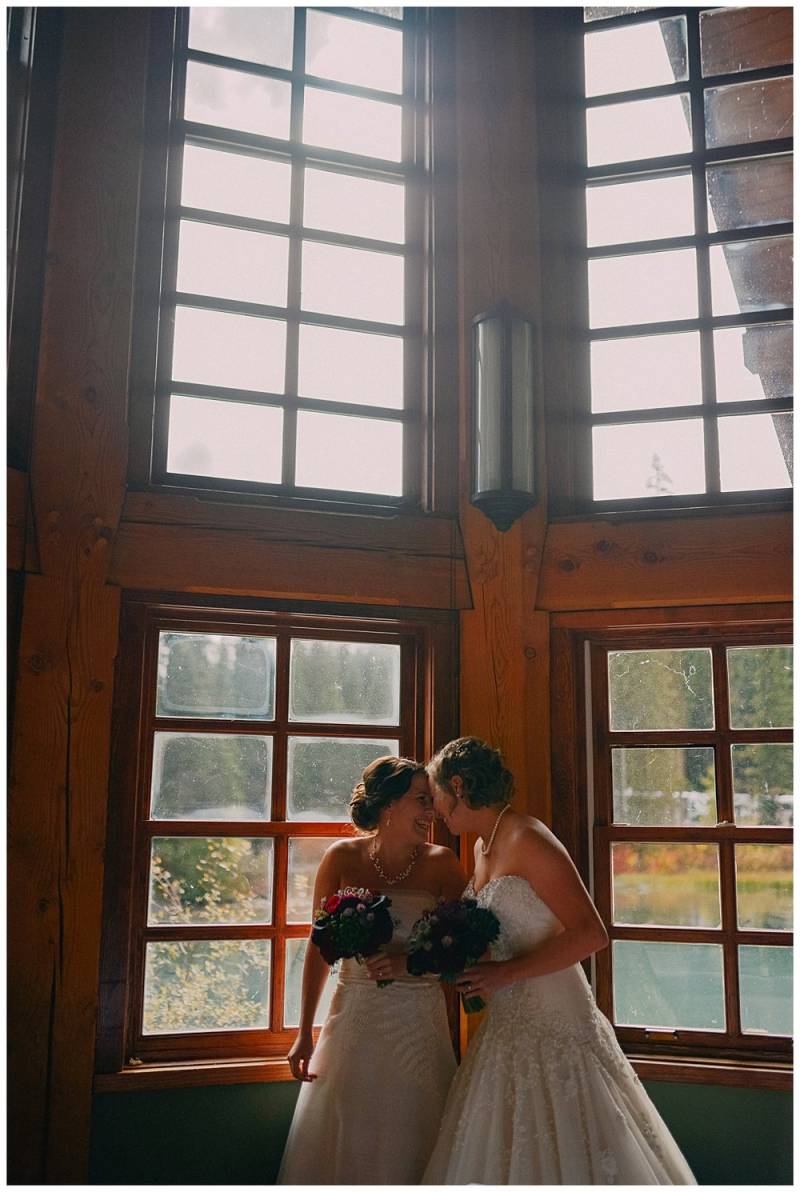 Brides in white dresses touch foreheads holding bouquets in naturally lit cabin