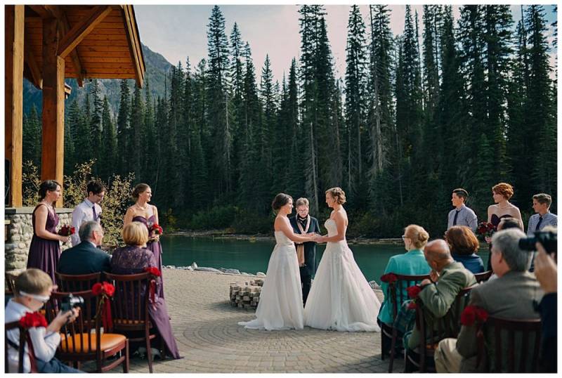 Brides in white dresses holding hands beside cabin and blue river with tall trees in background
