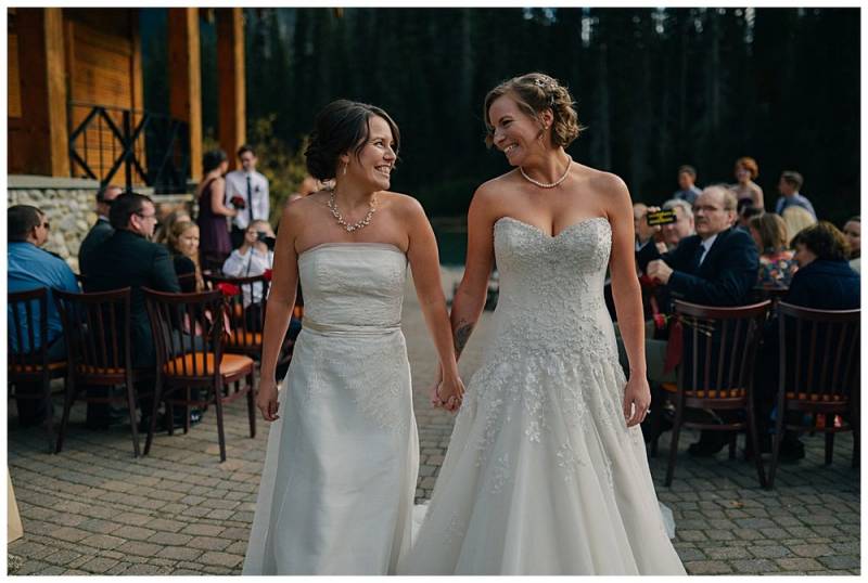 Brides walking down aisle smiling at each other in white dresses 