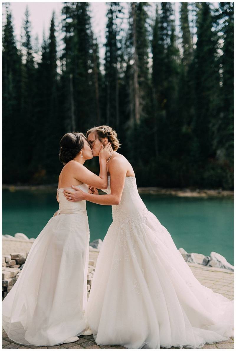 Brides kiss wearing white dresses at the edge of teal water and forest