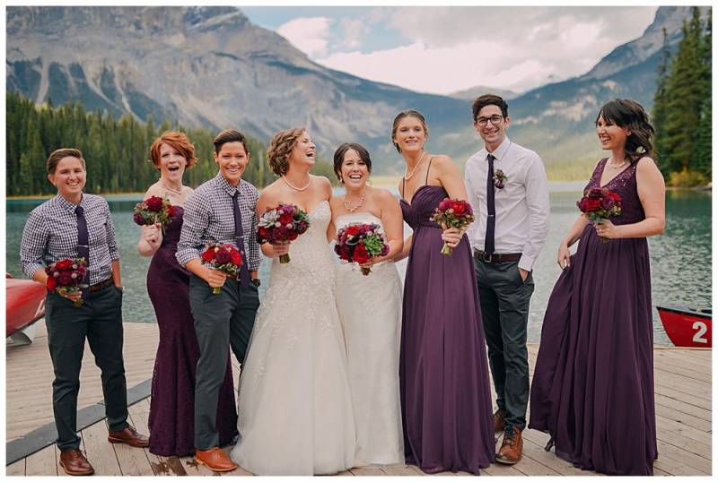 Bridesmaids and groomsmen stand on dock holding red bouquets wearing white and burgundy dresses