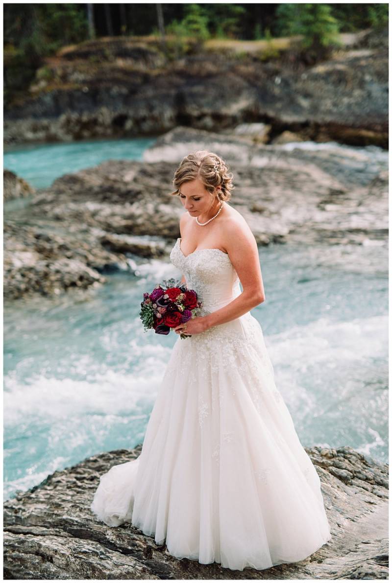 Bride in white dress holding red bouquet stands on rocks on river looking down