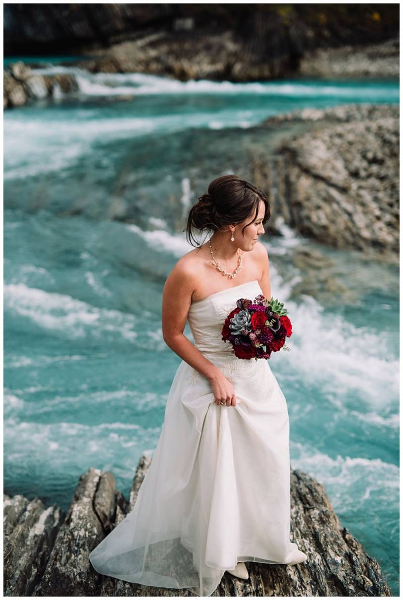 Bride in white dress holding red bouquet standing on rock on river  