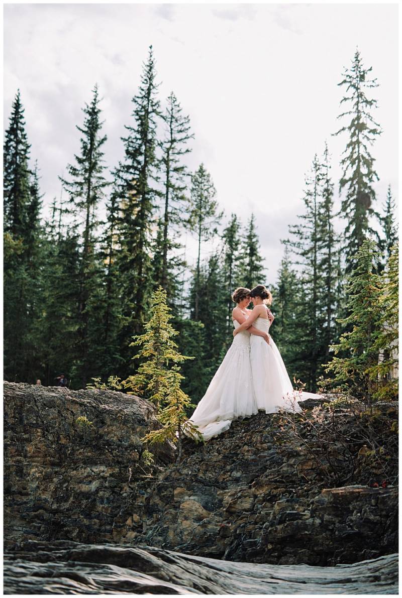 Brides in white dresses embrace on rocky riverbank with forest background