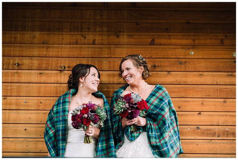 Brides wearing green plaid shawl holding red bouquets laughing on light brown wood background