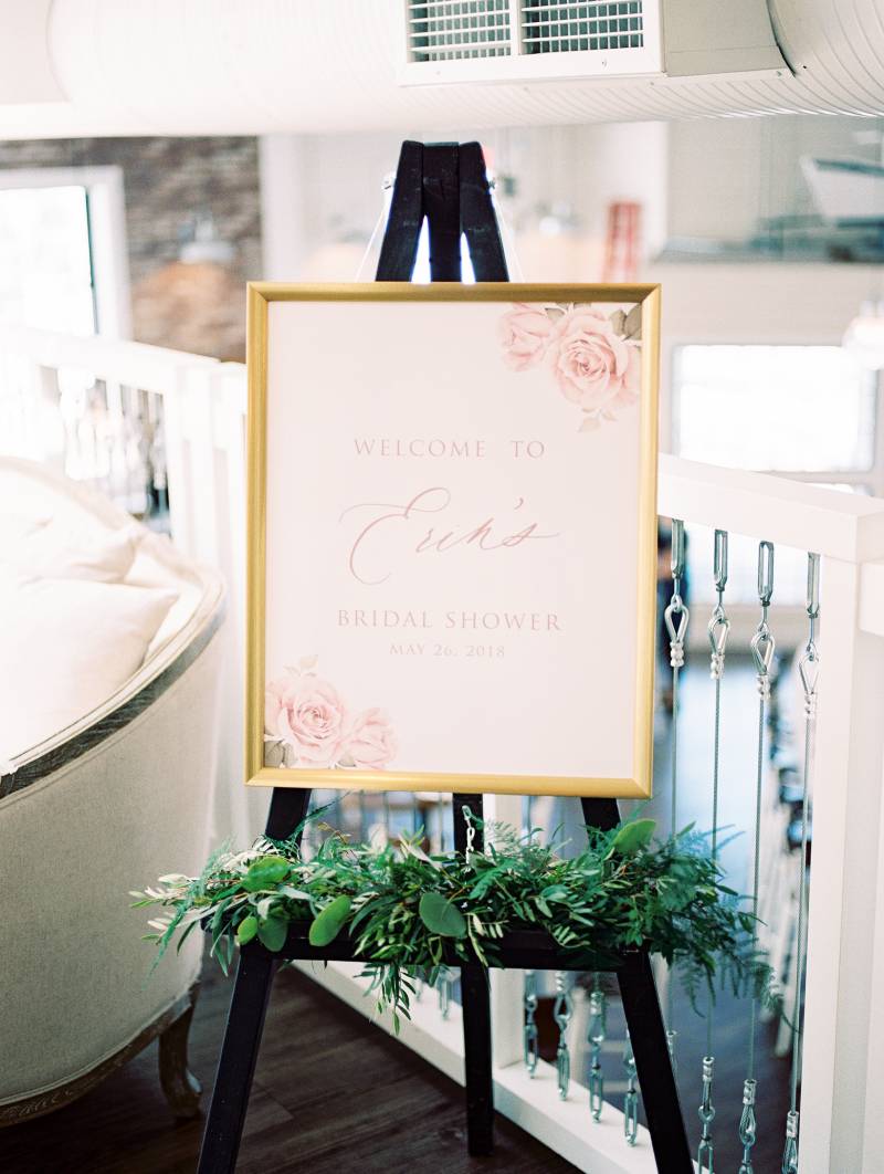 Pale pink bridal shower sign on black easel with greenery accents