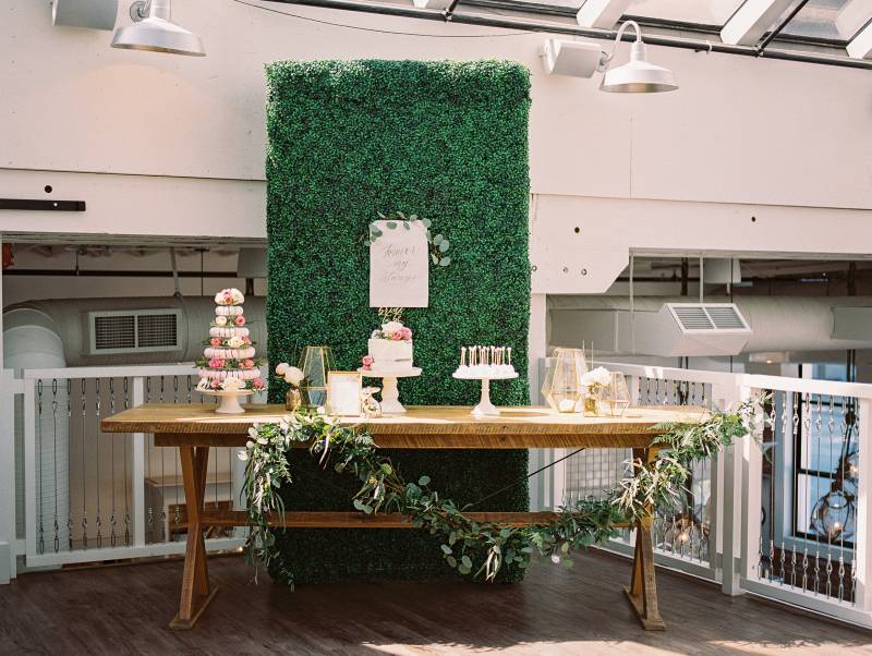 Large greenery wall and sign behind table with displayed cakes and candles with gold accents 