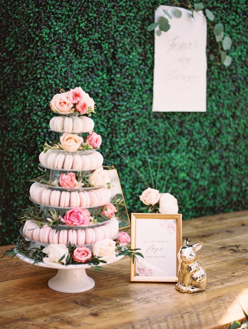 Greenery wall behind pale pink macaroon display with pink floral accents on table 