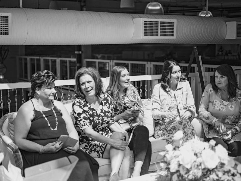 Five woman sitting in row on sofa laughing with floral arrangement in front