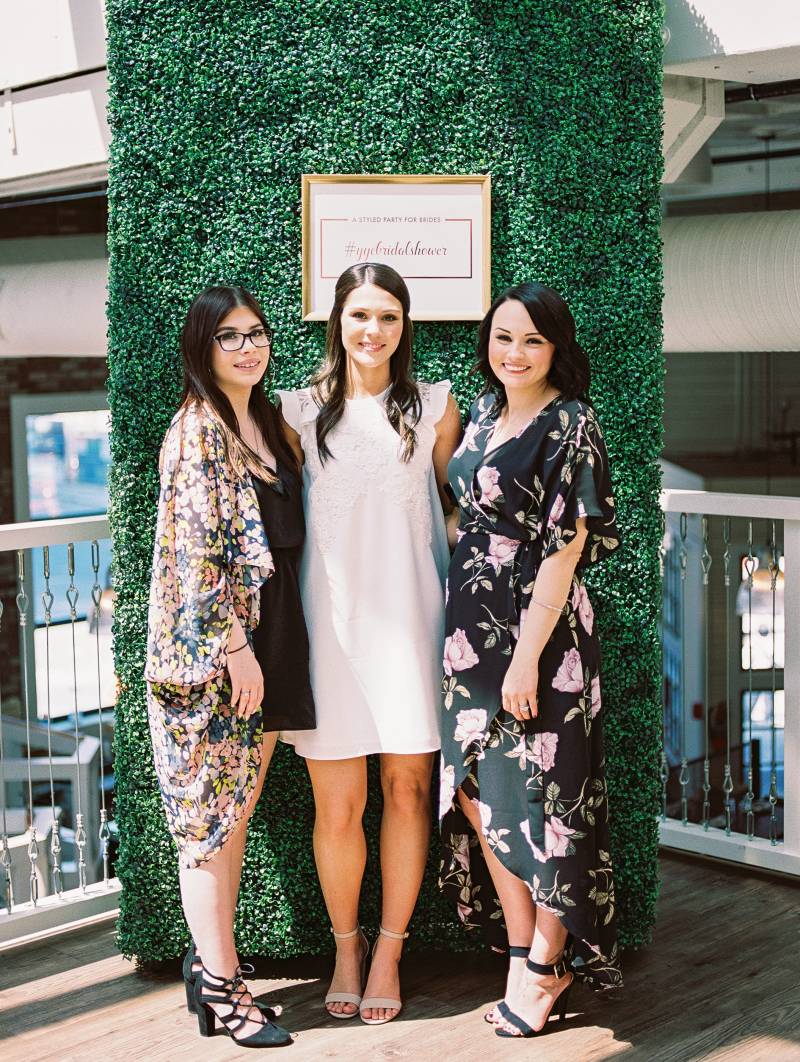 Three woman stand together in white and floral print dresses in front of large greenery wall 