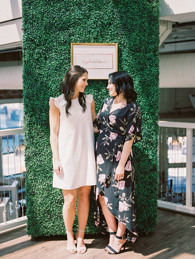 Two women in white and floral dresses stand together looking towards each other in front of large greenery wall 