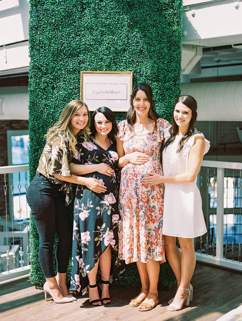 Four women standing together touching belly of pregnant women in front of greenery wall
