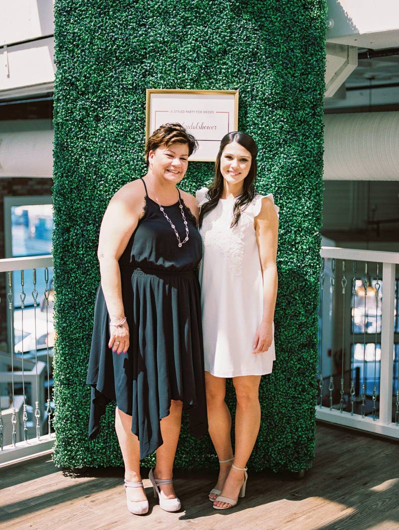 Two women one in black dress and one in white smiling in front of large greenery wall 
