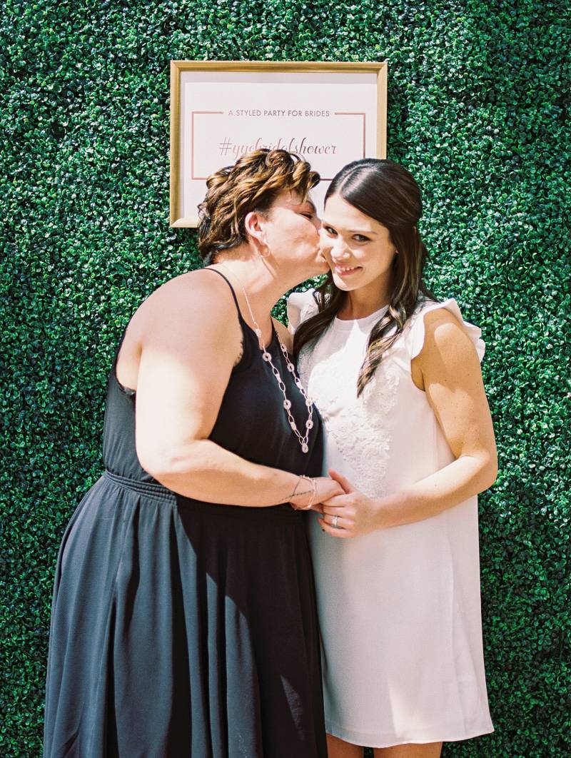 Woman in black dress kissed woman in white dress on cheek in front of greenery wall